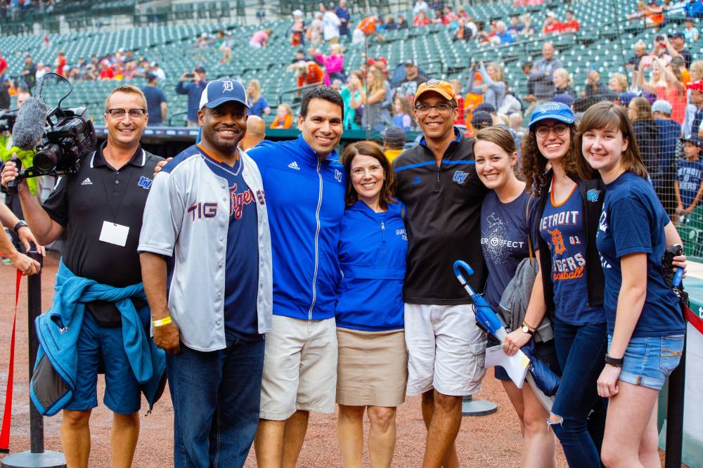 A group of the gvsu alumni staff and posing for a photo while standing on the infield of comerica park. There is also a camera man